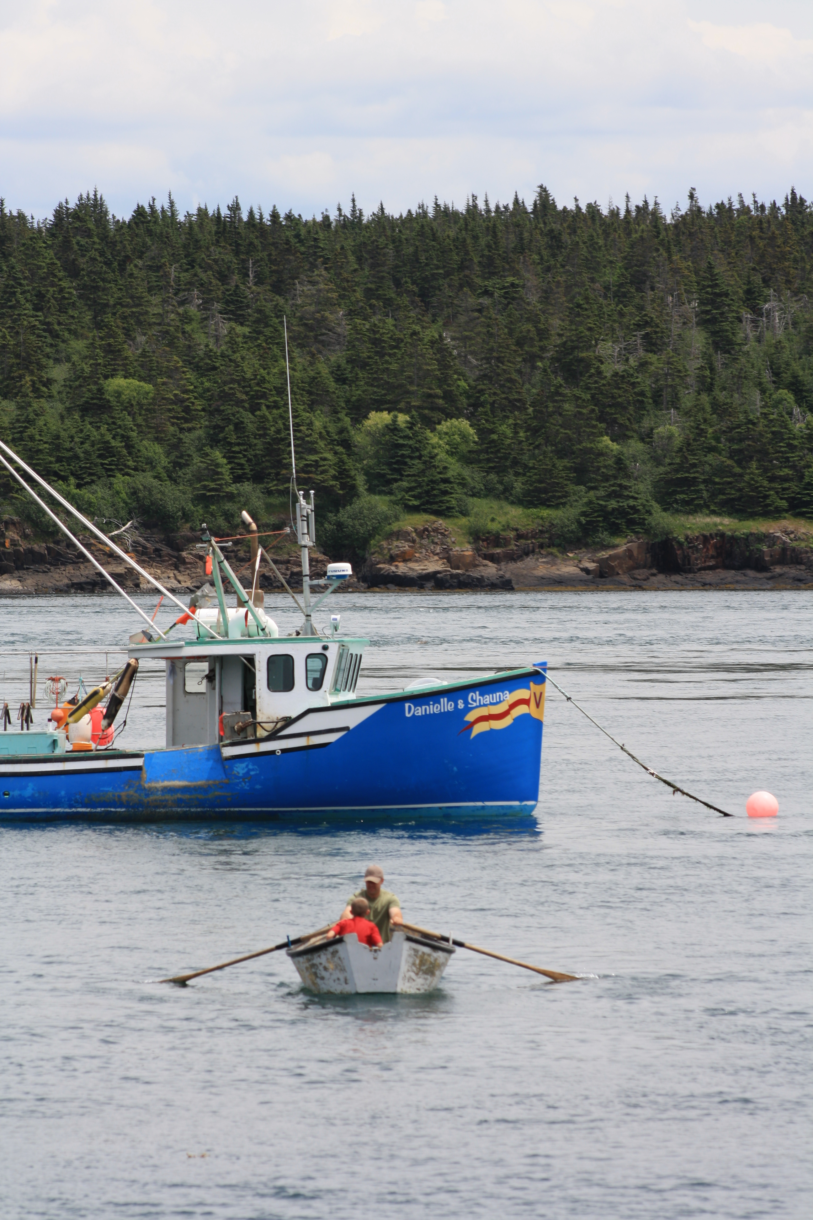 A man and his son row a small boat towards a larger fishing vessel. Behind the vessel is a forest coastline.