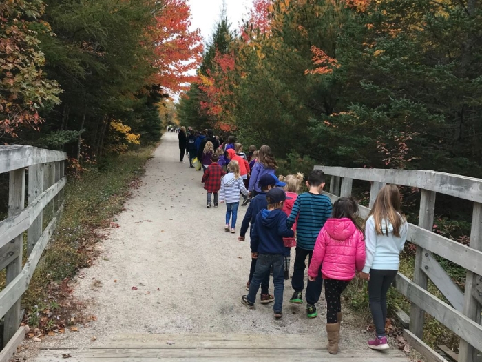 a group of school aged children walking on a gravel path with wooden railings on the side of a section of it. trees, with their leaves showing fall colours, are on either side of the path