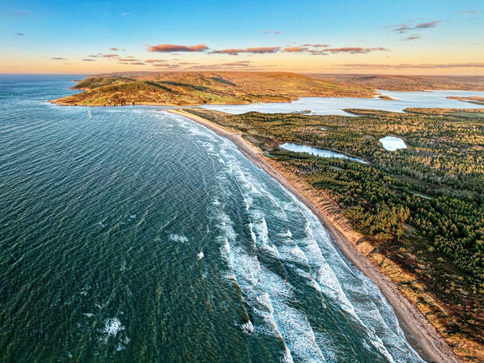 Ariel view of West Mabou Beach with a rich greenish blue ocean on the left and on the other side a sand beach with green grass on the right hand side.